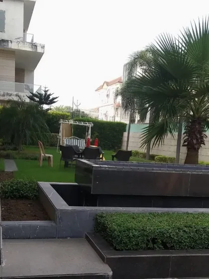 A view of a private residence in Panipat, showing a well-maintained lawn with a seating area in the background. The foreground features a modern tiered water feature and neatly trimmed hedges.