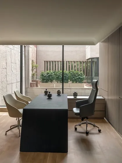 A meeting space within a director's cabin at 'The Layered Office'. The sleek, modern desk and chairs are positioned to take advantage of the view into the private courtyard, blending indoor and outdoor environments.