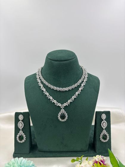 A graceful double-layered AD necklace. The top layer is a simple floral chain, while the bottom layer features a pear-shaped pendant. It is available in white, pink, green, and blue stones.