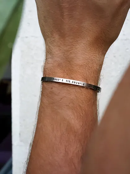 A close-up view of a silver cuff on a man's wrist, clearly showing the detailed engraving of latitude and longitude coordinates.