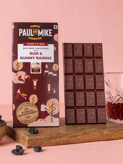 A moody shot of a rum and raisin dark chocolate bar, styled with a glass of rum. This pairing immediately tells a story about the product's rich, adult flavour.