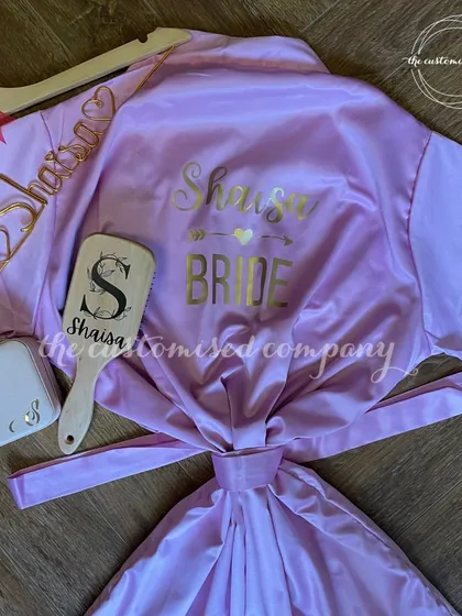 A curated collection of custom bride essentials to add a touch of magic to the wedding journey. This set includes a personalized lilac satin robe, a custom hanger, a jewelry box, and a matching hairbrush.