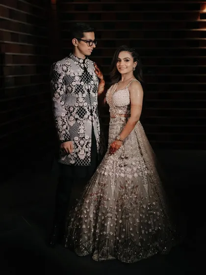 A classic, full-length portrait of the couple in their elegant Sangeet attire against a dark, textured wall.