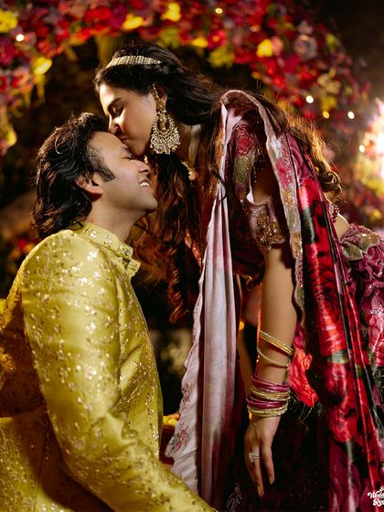 A tender kiss on the forehead under a beautifully decorated floral arch. A romantic portrait from a vibrant Sangeet night.