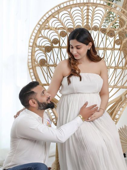 A sweet moment between expecting parents. The partner kneels to connect with the baby bump, set against our signature boho backdrop with a peacock chair and pampas grass.
