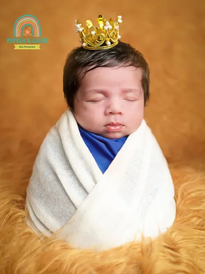 A close-up of a newborn prince wearing a crown, swaddled in white against a warm, furry backdrop. This highlights the delicate features of the baby.