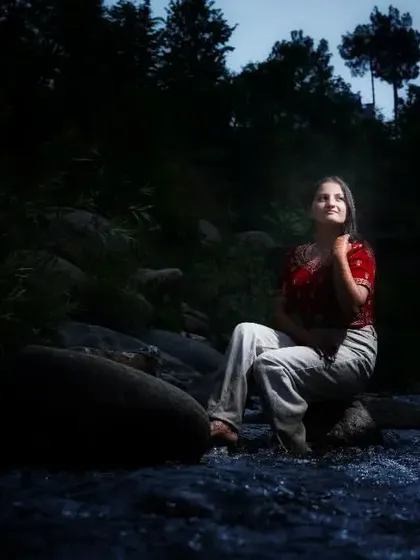 A dramatic portrait of a woman sitting by a river at dusk. The creative lighting highlights her against the dark, natural surroundings, creating a moody and contemplative lifestyle photograph.