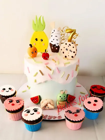A playful cake and cupcake set with cute fruit and cookie characters. The main cake is topped with a pineapple, ice cream cone, and cookie, surrounded by expressive cupcakes.