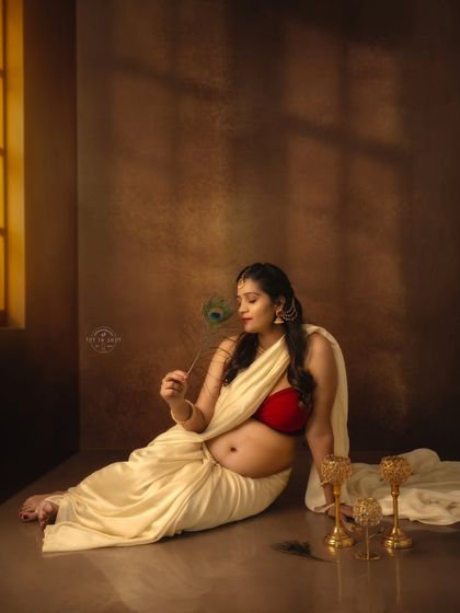 A quiet, contemplative moment captured in a low-key setting. The mom-to-be is seated, holding a peacock feather, with beautiful light coming from a window.