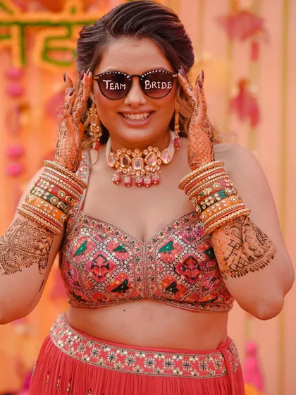 The bride is all smiles, showing off her "Team Bride" sunglasses during her Mehendi ceremony. This is a fun, candid shot that captures the bride's personality and the festive mood.