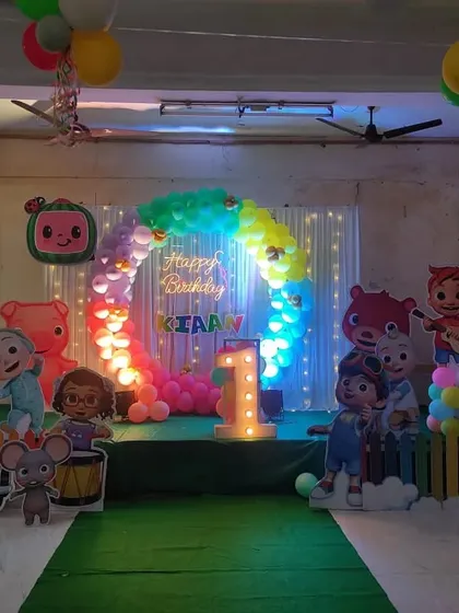 This Cocomelon party setup features a full stage with a green turf floor, a rainbow balloon arch, and cutouts of all the favorite characters, making it a perfect play area for the little ones.