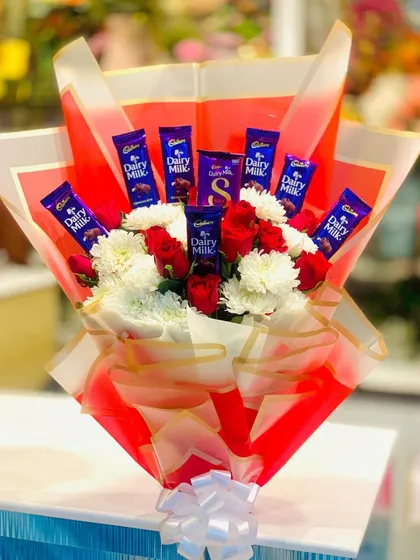 A vibrant bouquet combining fresh flowers with chocolates. This design features red roses and white chrysanthemums surrounded by bars of Cadbury Dairy Milk, all wrapped in bright red paper.