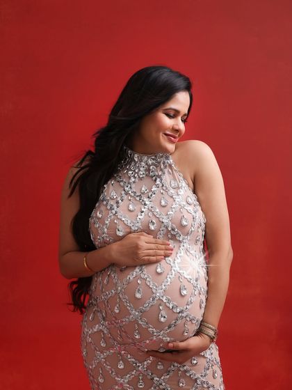 A close-up portrait that captures the radiant smile and gentle glow of the mom-to-be, her sparkling gown adding a touch of glamour against the bold red backdrop.