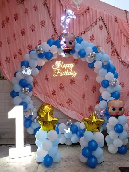 A Boss Baby themed first birthday party. This setup features a blue and white balloon arch, character foils, and a large '1' marquee light.