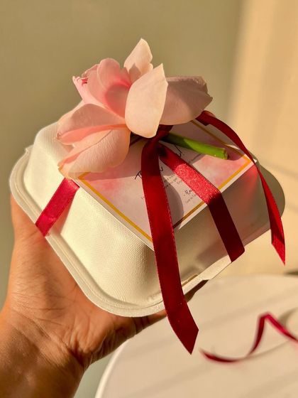 A close-up of a bento box gift-wrapped with a red ribbon and a delicate pink rose, held to show its giftable size and presentation.