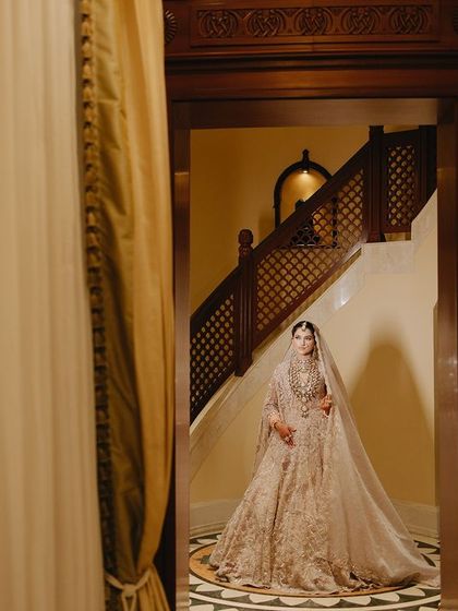 A wide shot of Megha in a grand hallway, her long veil trailing behind her. This image captures the scale and elegance of her bridal look within the luxurious venue.