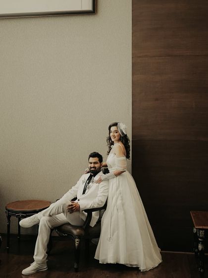 A stylish, posed portrait with a classic feel. The groom sits while his bride stands beside him, a timeless composition that highlights their elegant white wedding outfits.
