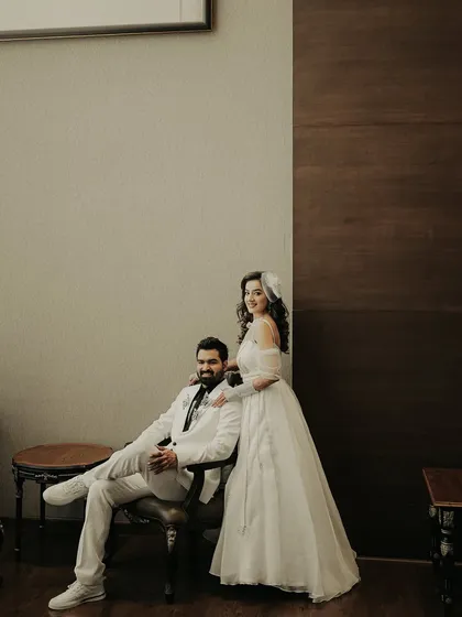 A stylish, posed portrait with a classic feel. The groom sits while his bride stands beside him, a timeless composition that highlights their elegant white wedding outfits.