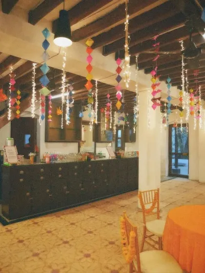 An indoor Mehendi setup with colorful paper hangings and fairy lights. The space is designed to be festive and fun, with a dedicated bar area for guests to enjoy.