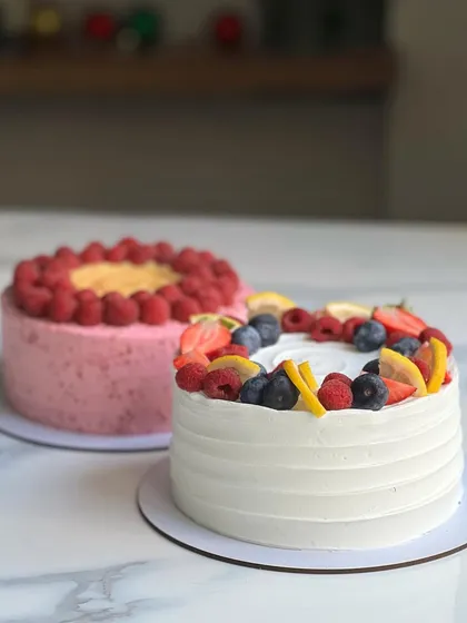 A beautiful duo of berry cakes. One is a Lemon Raspberry cake with a lemon curd center, and the other is a fresh fruit cake topped with a mix of berries and citrus.