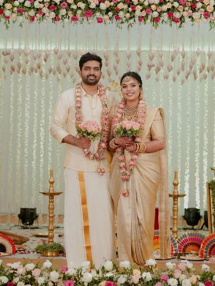 A classic portrait of the bride and groom at their mandap, holding bouquets and looking elegant in their matching cream and gold attire.