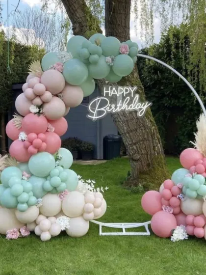 A beautiful outdoor birthday setup in a garden. The circular arch is decorated with pastel green and pink balloons and accented with pampas grass for a soft, natural feel.