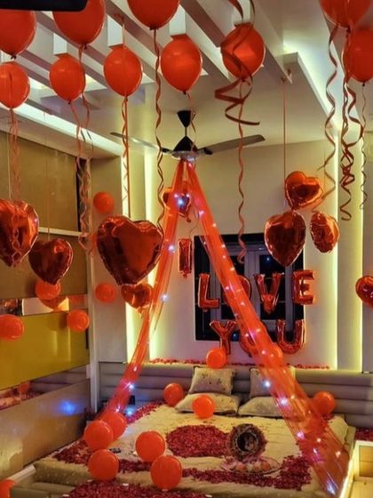 A passionate and romantic room setup entirely in red. The bed is decorated with a canopy, fairy lights, and rose petals, while red heart-shaped and latex balloons fill the room, creating an atmosphere of pure love.