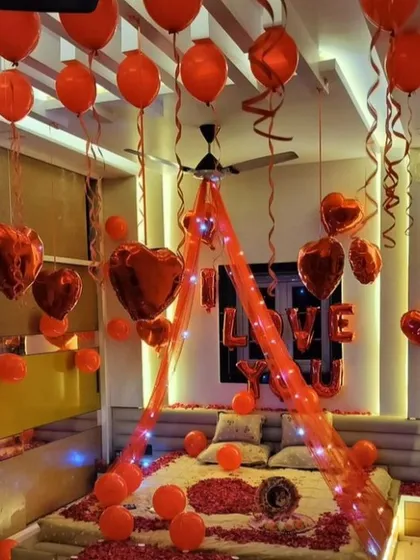 A passionate and romantic room setup entirely in red. The bed is decorated with a canopy, fairy lights, and rose petals, while red heart-shaped and latex balloons fill the room, creating an atmosphere of pure love.