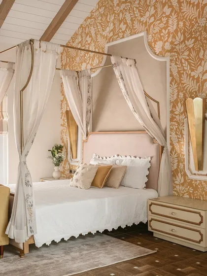 A bedroom from our DP Debut showcase, featuring a romantic four-poster bed with a tall, shaped headboard and delicate drapery. The mustard yellow floral wallpaper adds a touch of vintage charm.