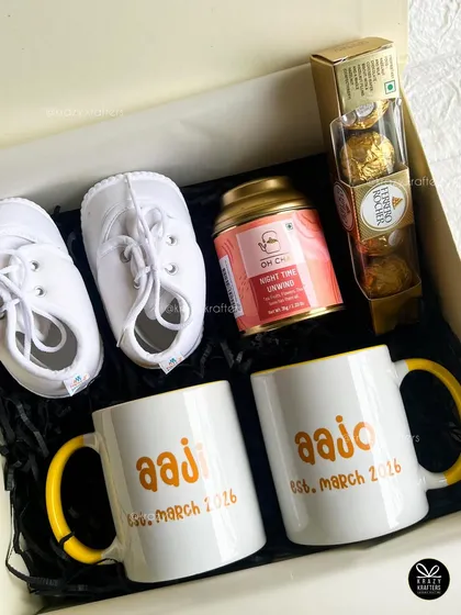 This 'grandparents-to-be' hamper includes personalized 'Aaji' and 'Aajo' mugs, tiny white baby shoes, Ferrero Rocher chocolates, and a tin of relaxing tea.