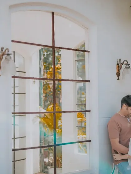 An intimate portrait of a couple against a large, elegant window. The natural light and surrounding foliage create a soft and romantic mood for this pre-wedding photo.