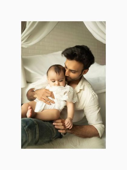 A father gently kisses his baby daughter's head in this close-up studio portrait. The soft lighting and white fabrics create a feeling of warmth, safety, and deep affection.