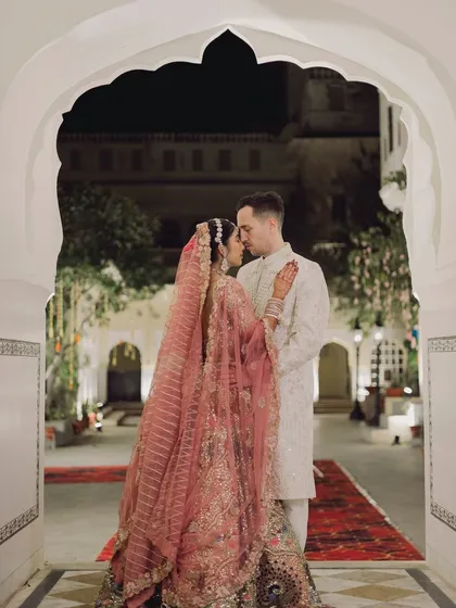 A romantic couple portrait framed by a classic scalloped archway. The bride's intricate pink lehenga and the groom's elegant sherwani are beautifully captured in this timeless shot.