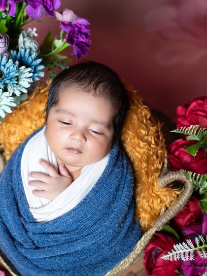 A newborn swaddled in blue, sleeping in a basket surrounded by a beautiful arrangement of flowers.