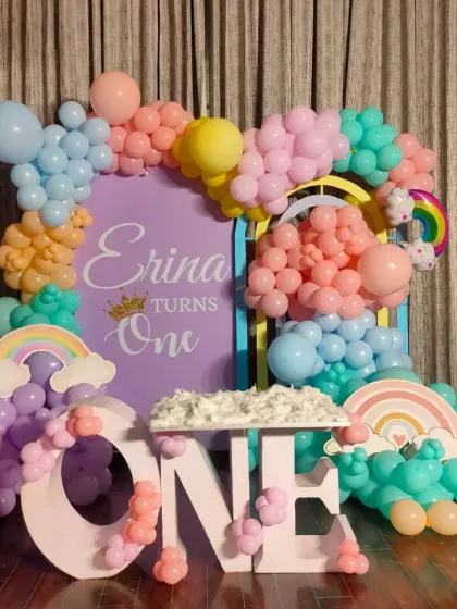 Even a small space at home can be magical. Here is a pastel rainbow theme for a first birthday, featuring a custom backdrop for "Erina turns One" and large "ONE" marquee letters decorated with matching balloons.