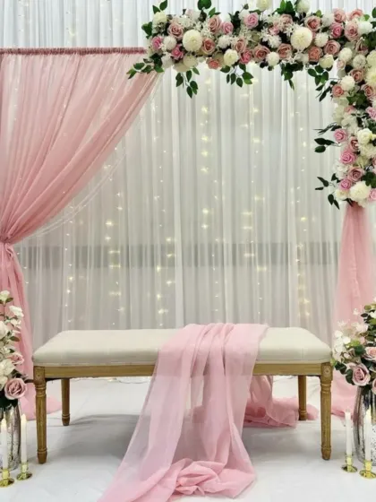 An elegant backdrop for an engagement or bridal shower. The setup features a sheer pink drape over a fairy light curtain, framed by an arch of beautiful artificial flowers.