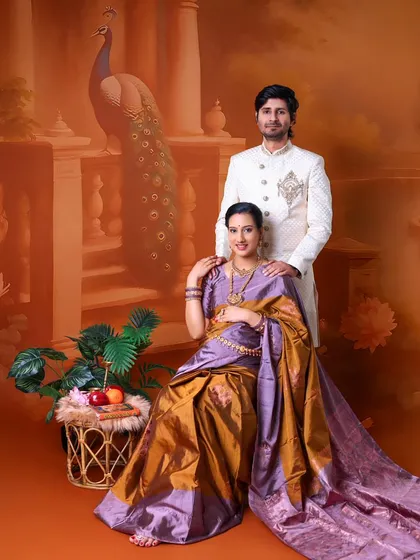 A regal portrait of the expecting couple. The mom-to-be is seated, draped in a beautiful saree, while her partner stands by her side, set against a traditional painted backdrop.