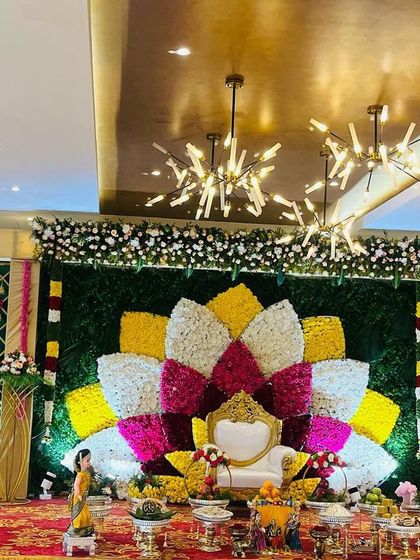 A wide-angle view of a magnificent Seemantham stage. The decor features a multi-colored floral lotus, decorative side panels, and a beautifully arranged display of traditional offerings.
