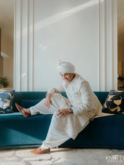 A relaxed portrait of the groom in his white wedding attire, seated on a modern sofa. This candid shot captures a moment of ease and happiness on his wedding day.