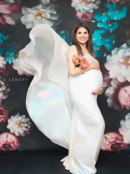 A fine art maternity portrait against a rich floral backdrop. The flowing white fabric of the gown adds a touch of drama and elegance, creating a timeless, painterly photograph to celebrate your pregnancy.