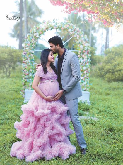 A romantic outdoor portrait of a couple under a floral arch. The soft pink gown and the husband's gentle kiss create a fairytale-like moment.