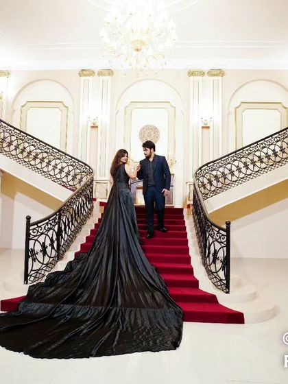 A wide shot of a couple on a grand staircase with a red carpet, her black trail gown spread out, creating a dramatic and cinematic entrance scene.