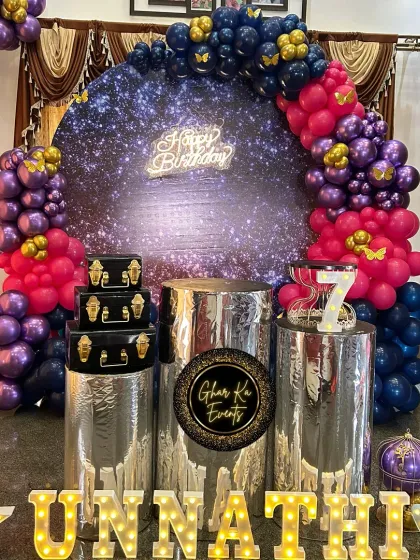 A galaxy-themed birthday party with a sparkling backdrop. The decor features a deep purple and pink balloon arch with gold butterfly accents, metallic silver plinths, and a 'Happy Birthday' neon sign.