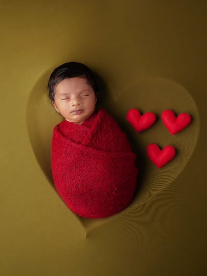 A newborn wrapped in red sleeps in a heart-shaped impression on an olive green background, with three small red hearts next to them.