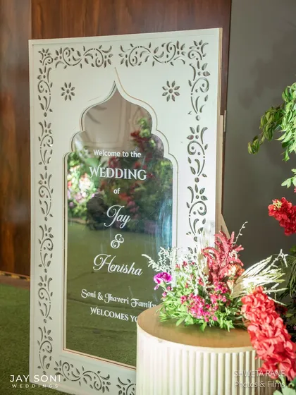 A close-up of the elegant mirrored welcome sign, framed with traditional Indian jali-cut work, reflecting the vibrant floral decor that surrounds it.
