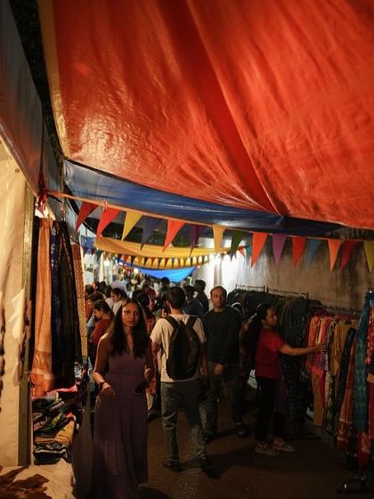 The evening crowd explores a narrow alley of stalls filled with textiles and clothing, under a colourful canopy of flags.