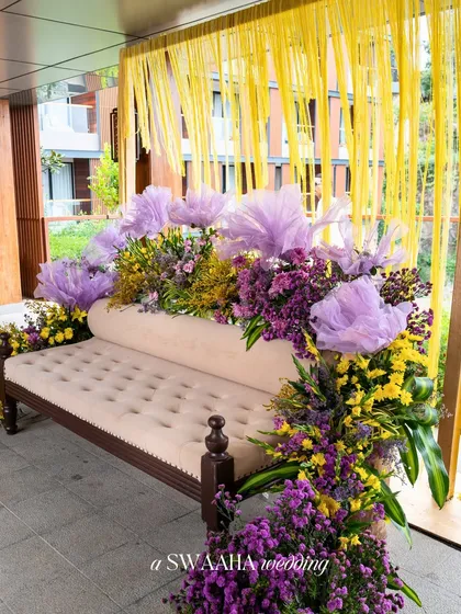 A close-up of a seating area, where a plush off-white diwan is surrounded by an explosion of purple and yellow flowers.