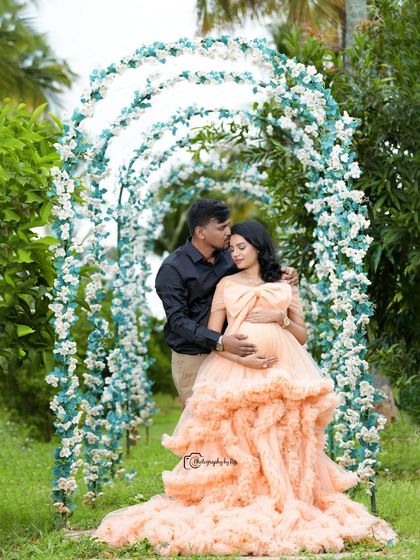 An intimate couple's portrait under a floral archway. The father-to-be's gentle kiss on the forehead captures the love and tenderness of the moment.