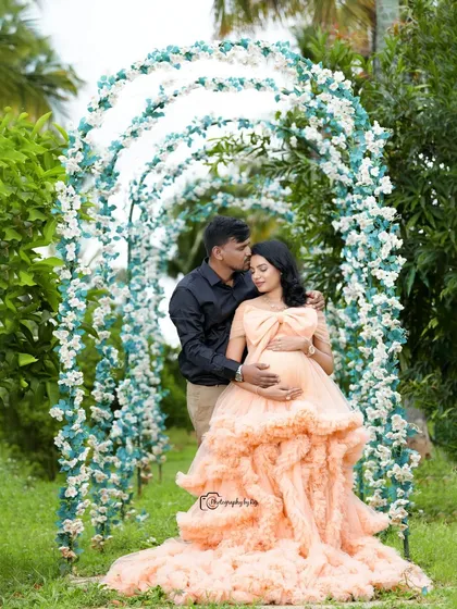 An intimate couple's portrait under a floral archway. The father-to-be's gentle kiss on the forehead captures the love and tenderness of the moment.