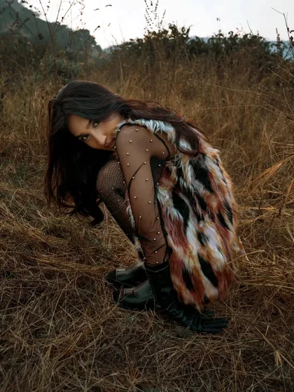 A fashion portrait taken in a field of tall, dry grass. The model's crouching pose and direct gaze create a sense of being wild and connected to the earth.